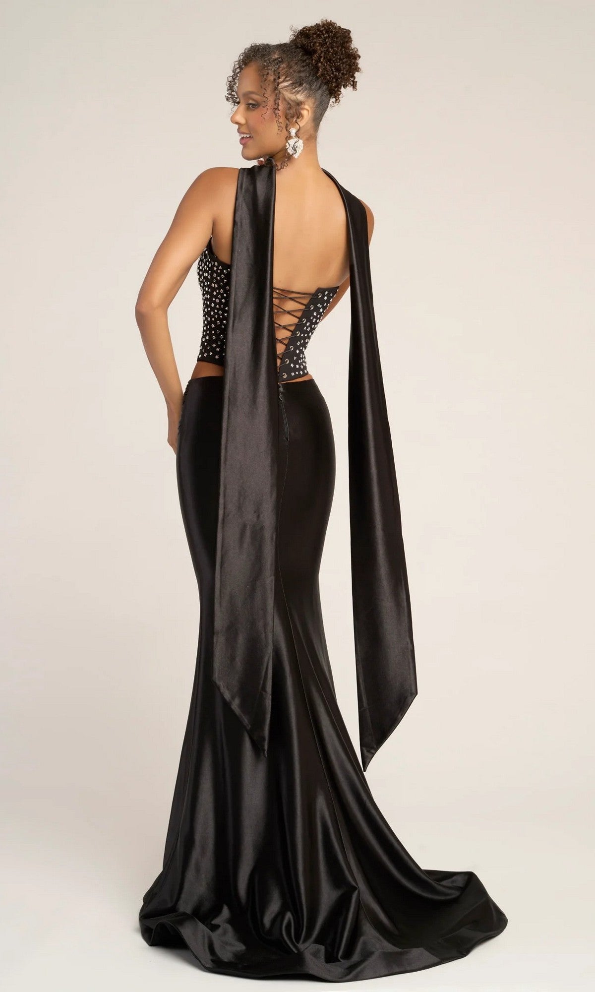 A woman wearing the Portia and Scarlett PS26042 long formal dress, featuring a black satin skirt, beaded corset top with lace-up back, and wide draped straps, stands looking over her shoulder.
